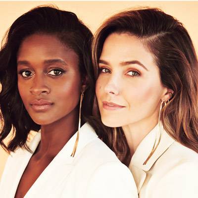 Portrait of Sophia Bush and Nia Batts- First Women's Bank Strategic Advisors, in stylish white suits, standing close together. Both wear long earrings, adding elegance to their attire.