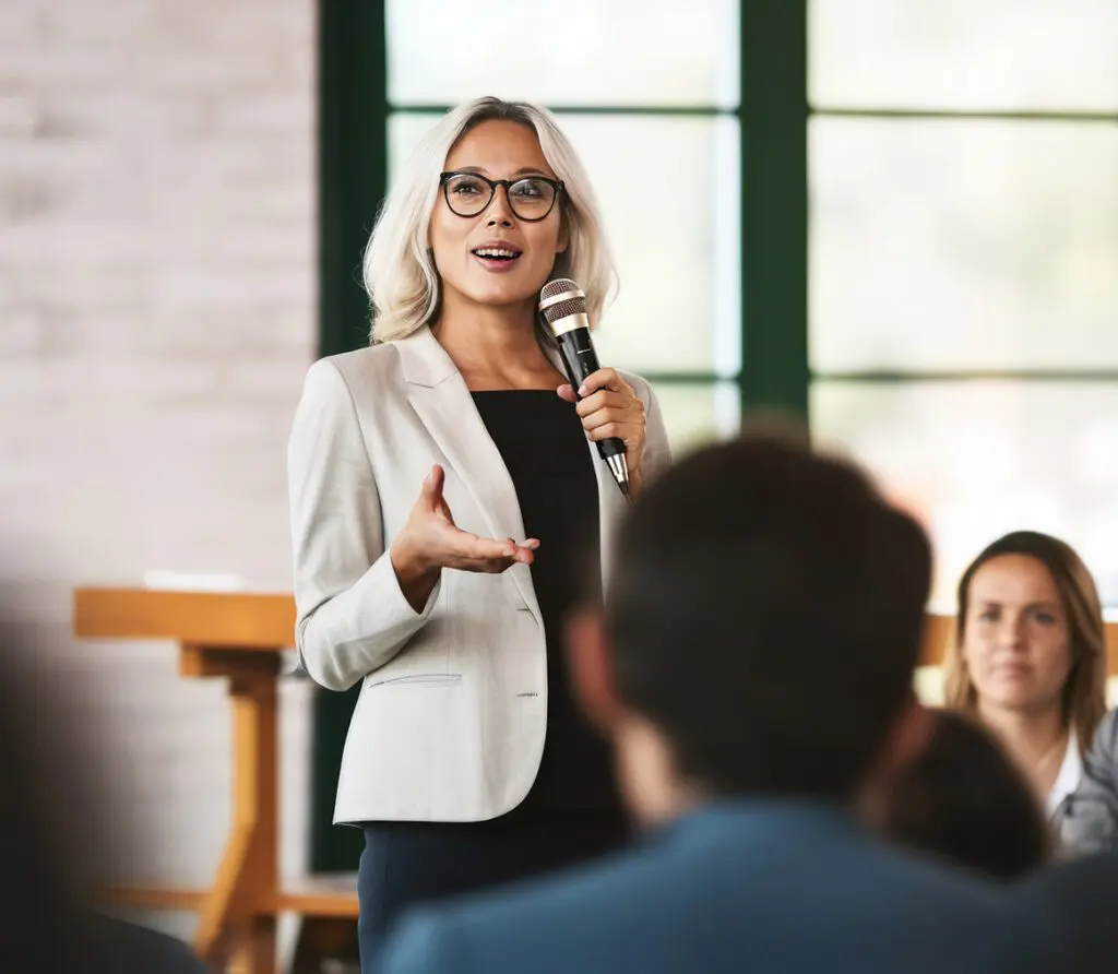 A confident woman with blonde hair delivers a speech at a leadership retreat. She exudes professionalism in a suit, complemented by stylish round black eyeglasses, emanating expertise in her presentation.